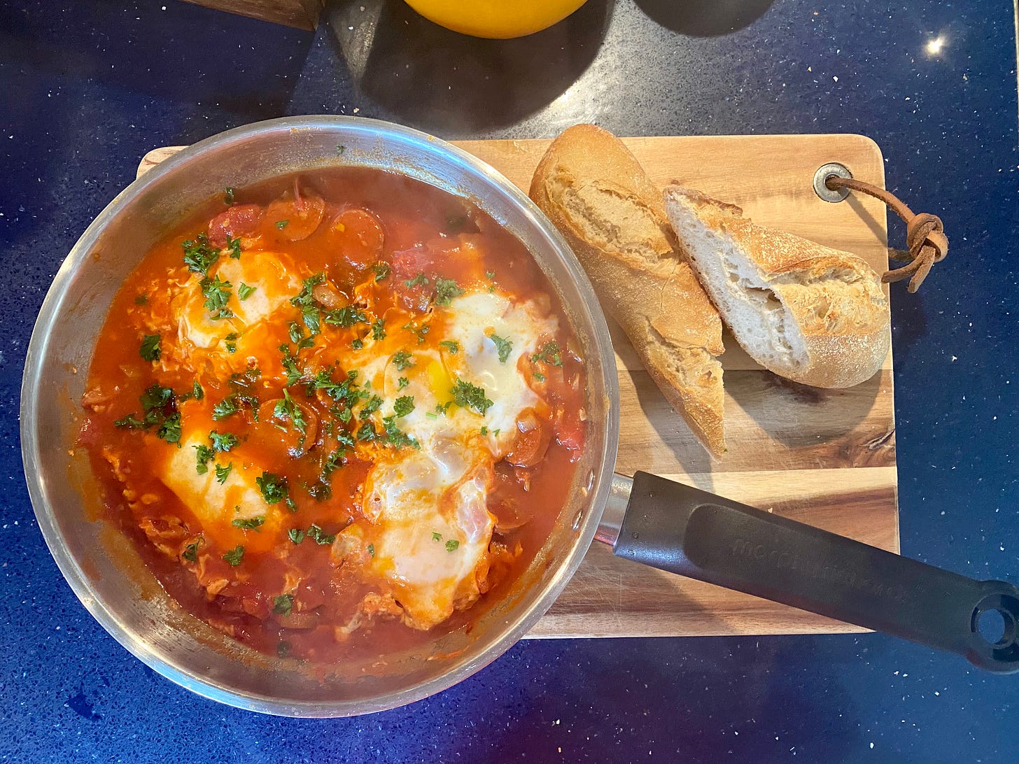 blue worktop, with a wooden board. On the board is a frying pan with a tomato-based mixture, eggs, and chopped green herbs. Some crusty bread is to the side. 