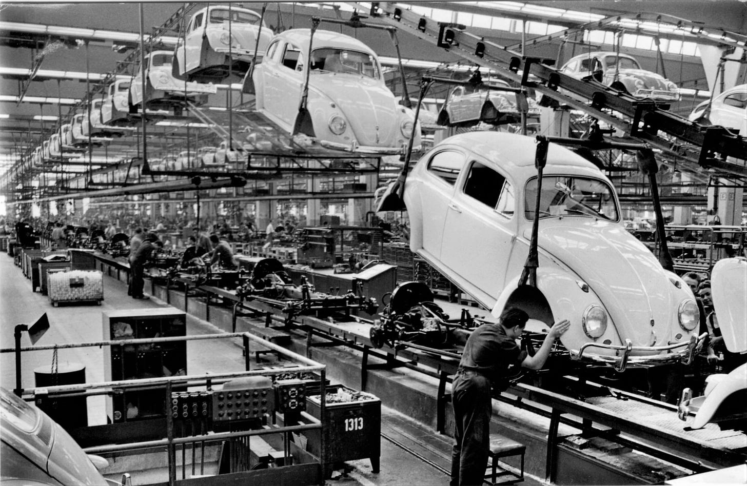 A black-and-white photo of a vintage Volkswagen Beetle factory asembly line.