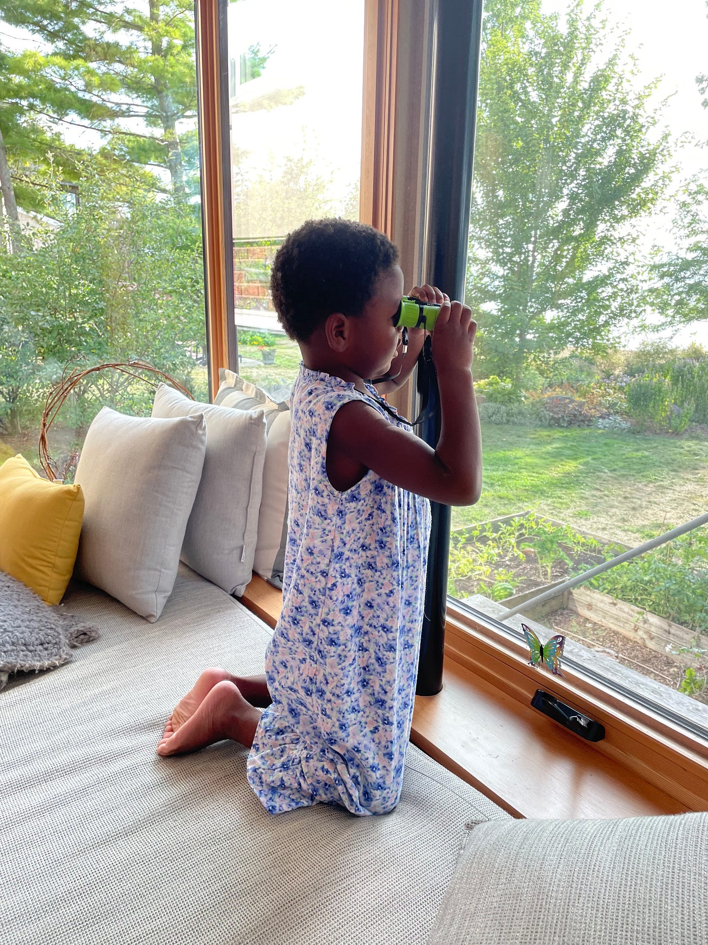 a Black kid in a long floral nightgown looks out the window with binoculars