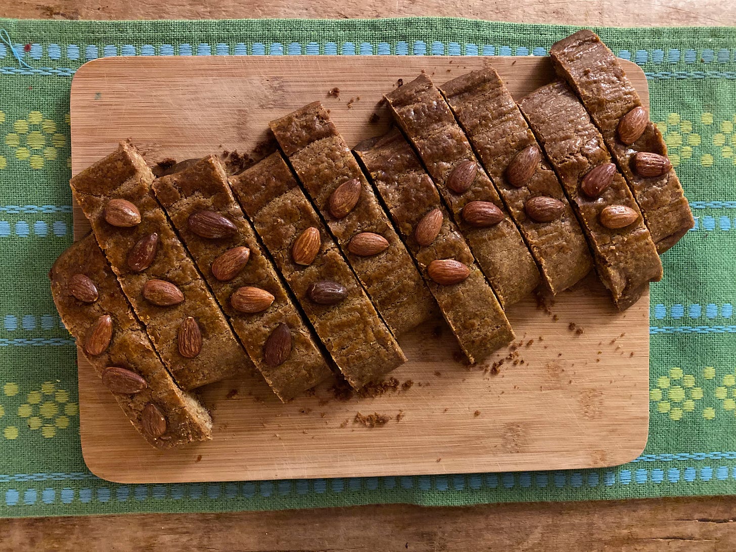 12 golden brown cookies, long oblong strips, sit on a cutting board on top of a green tablecloth. Each cookie has two almonds on top, and they are scrunched up next to each other; they’ve been cut from one large bake into twelve pieces.