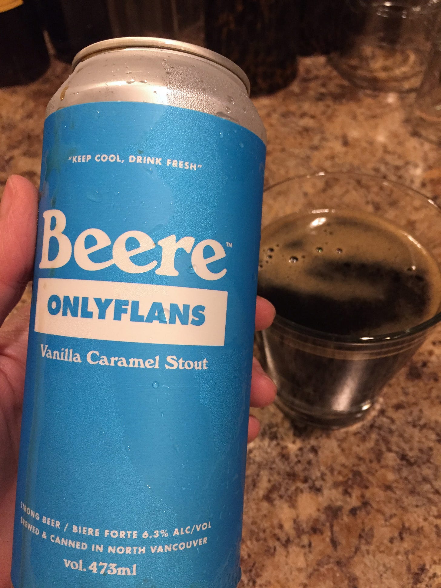 my hand holds up a blue-labelled beer can in the foreground, with a glass of dark beer in the background. The label reads, "Beere / onlyflans / vanilla caramel stout".