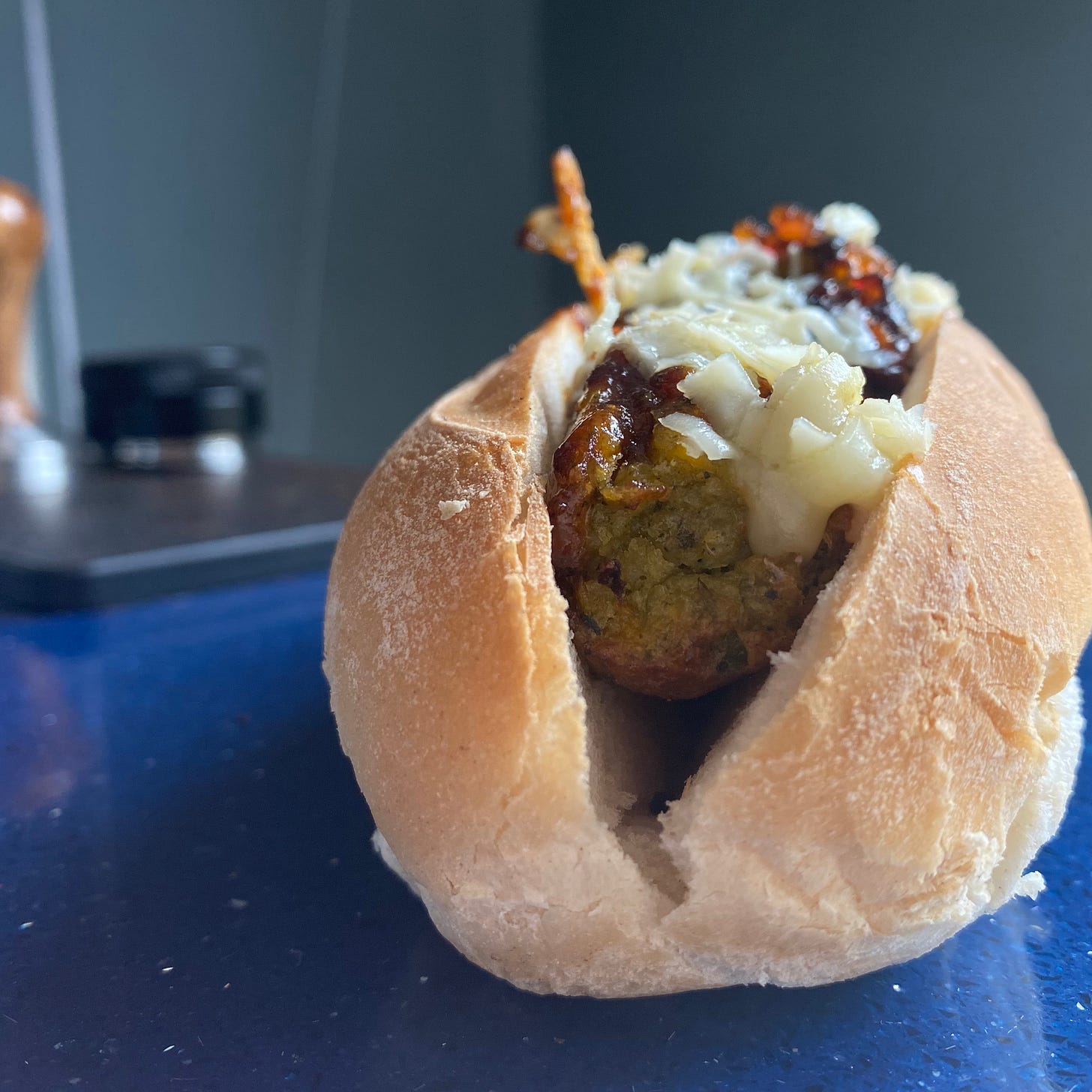 Hot dog roll filled with vegetarian sausage, and topped with onion chutney and grated cheese. Hot dog roll is placed on top of a blue quartz worktop, a grey wall behind.