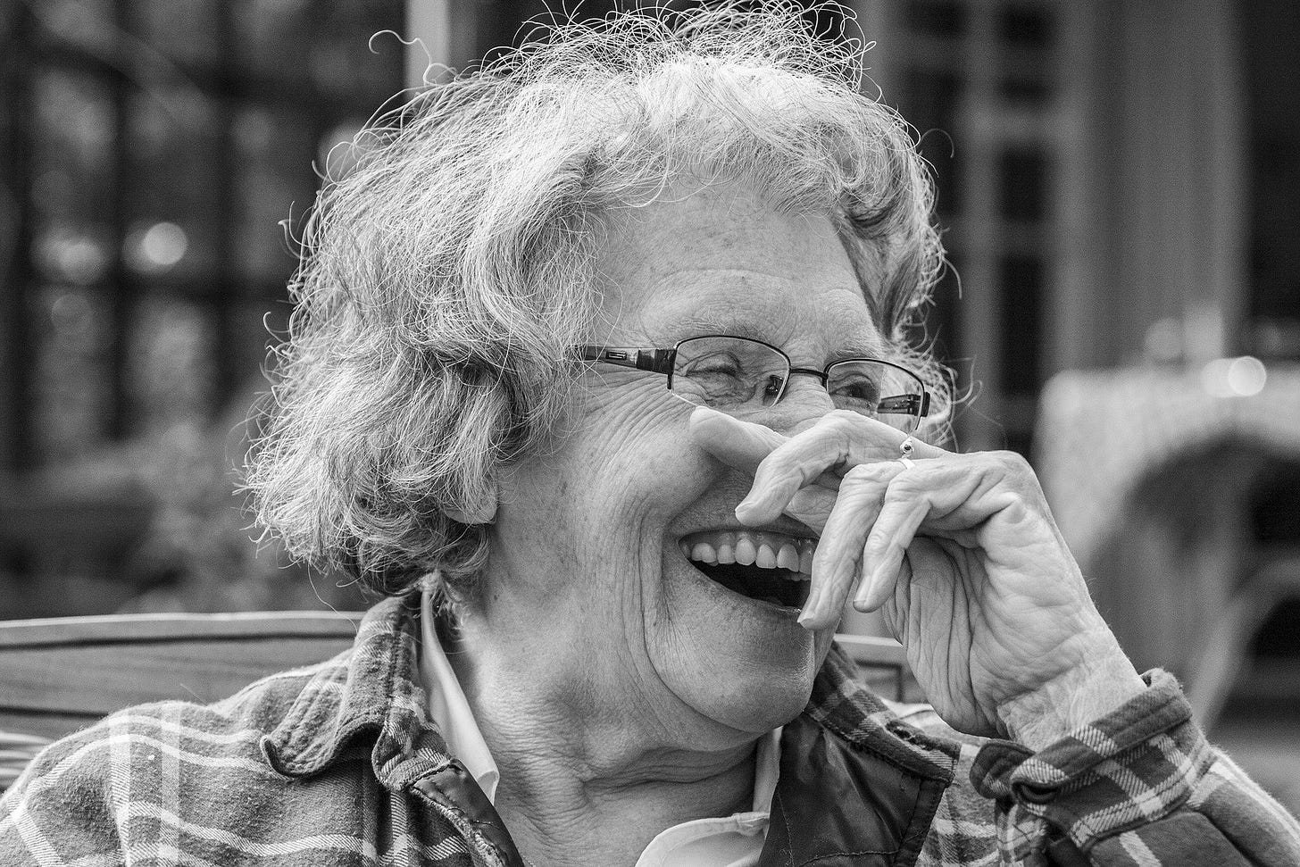 Black and white portrait photo of white woman with gray hair and glasses who is holding her right fist against her nose as she laughs, showing her false teeth.