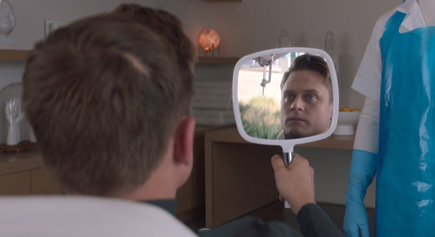 A white man with brown hair holds up a mirror to his face, in which a metal instrument prepares to insert a chip in his head.