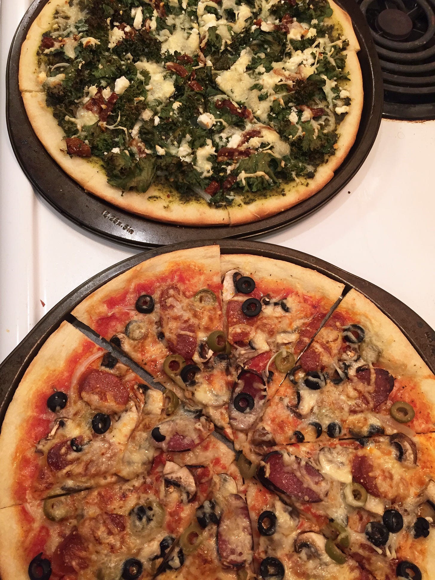Two pizzas in their pans on top of the stove. At the back, a pesto base with kale, ricotta, and sun-dried tomatoes. At the front, red sauce with salami, mushrooms, and olives.