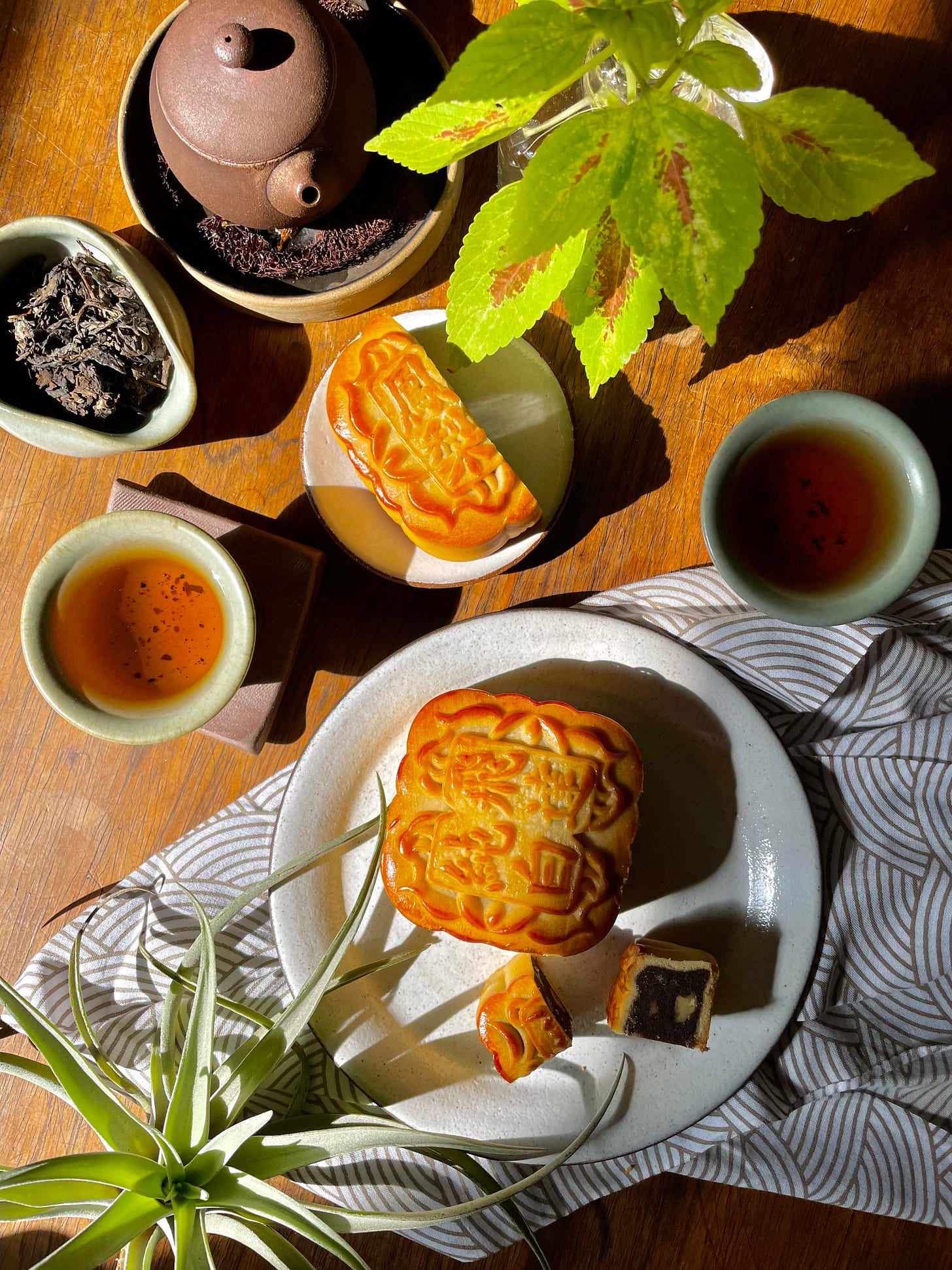 ID: A lovely sun drenched photo of a tea setup on a wooden table with cerulean ceramics, unglazed pottery, plants and decorative fabric.