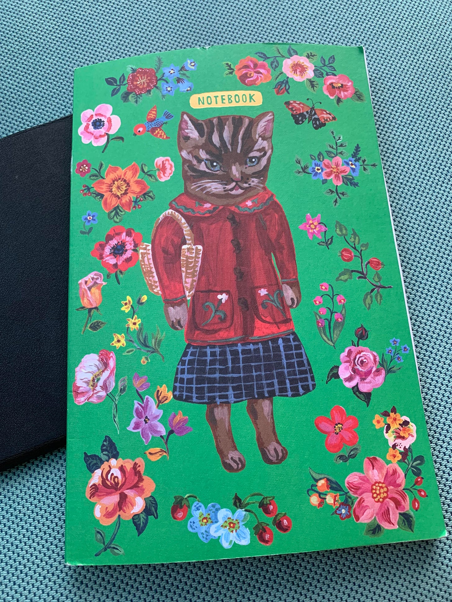 A green journal cover with an image of a brown cat wearing a red sweater and blue skirt and holding a basket, surrounded by flowers on a vibrant green background