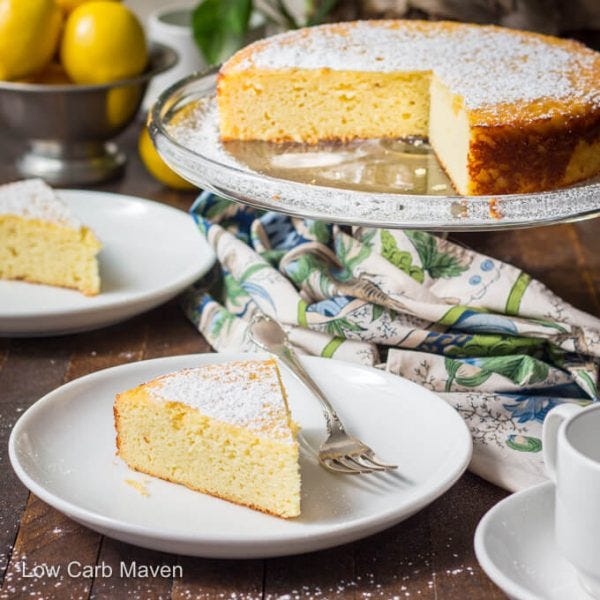 A flat yellow cake with icing sugar on top sits on a glass cake stand. A white and tea towel is spread under it, and below is a small white plate with a silver fork and a slice of the same yellow cake. The words 'Low Carb Maven' are across the bottom left corner