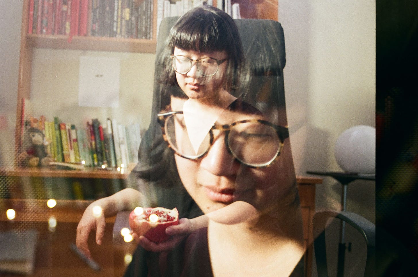A double exposure photograph, wherein two images are faded onto one another in a single image, of Jane sitting at her desk on a black office chair.  Jane is a genderfluid person with pale tan skin, with her eyes closed, wearing brown and black glasses and short black hair that slightly flares at the end with rounded bangs like a bowl. In one exposure Jane’s face is larger and closer to the camera; there are lights directly behind her. In the other, she is sitting on a black office chair and holding a sliced open half of a pomegranate. Behind her is a round, moon-like desk lamp. The desk behind her is a medium orange-brown and contains a head shelf, with books ordered in red, black, white, and the rest of the rainbow ordered on the top of the table. There’s a small brown monkey plushie near the left end of the desk. There’s a black and white ink illustration above the orange to blue books on top of the desk where there’s an open space below the desk shelves. Photo Credit: Divya Kaur (@soft.kaur on Instagram) 