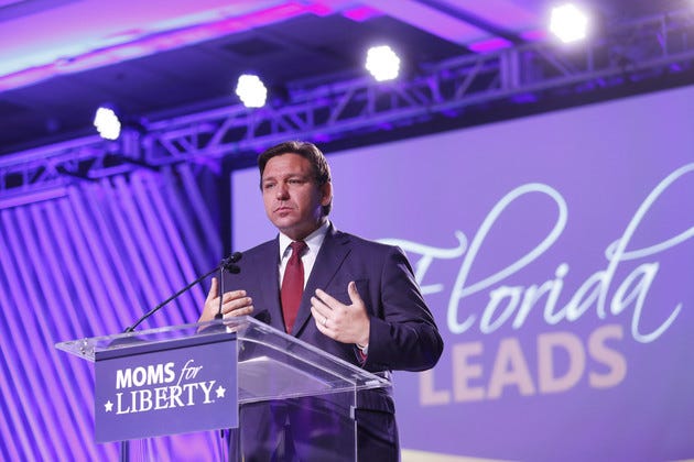 Ron DeSantis - a man wearing a blue suit and red tie - stands at a podium that reads Moms for Liberty