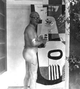 Le Corbusier is naked, standing side on in front of a vandalised wall, wearing glasses.