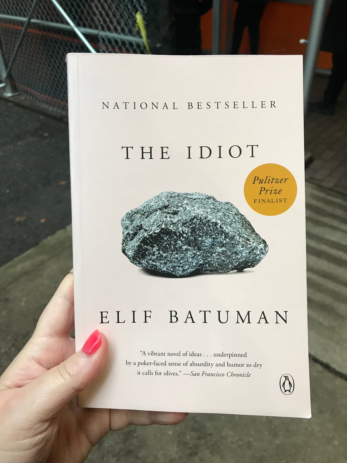 A hand holding a copy of Elif Batuman's The Idiot, with concrete and metal fencing behind the book.