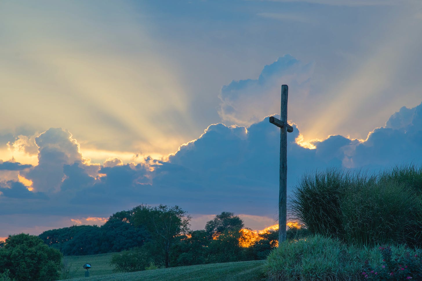 A wooden cross in a grassy field with the sun behind a cloud. 