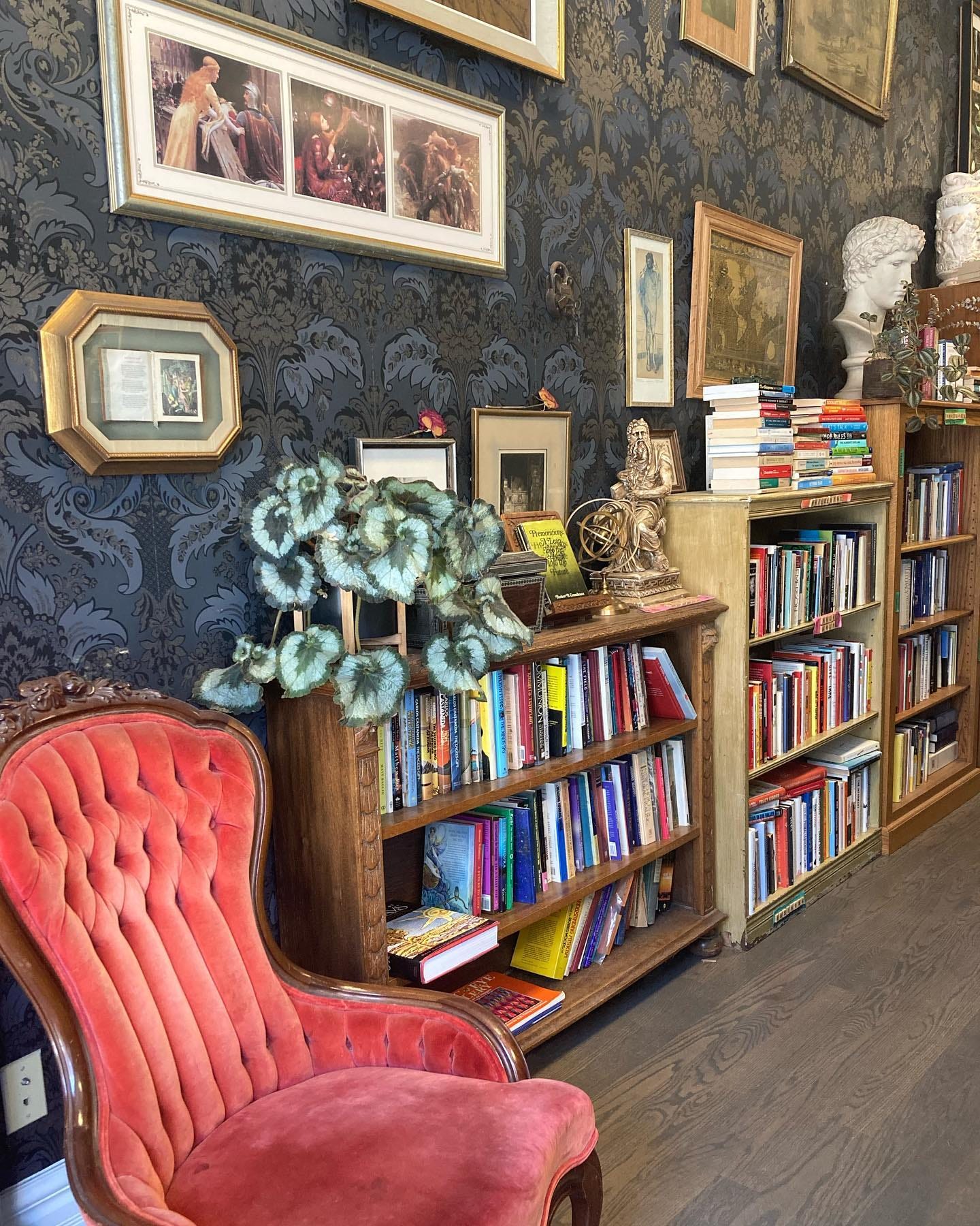 Antique orange velvet chair, bookcases ascending in height, ornate wallpaper in shades of grey-blue, with various pictures hung on it, plants and knickknacks