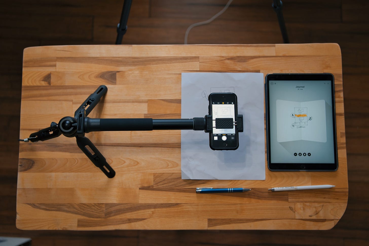 Overhead shot of tripod with extnesion holding an iPhone with the video camera app open, paper and pen below it, iPad pro and pencil beside it