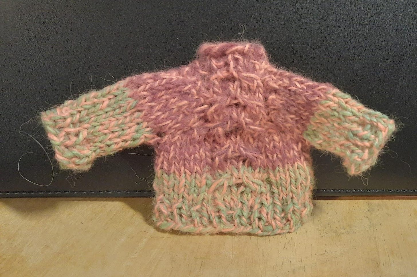 A small sweater in green and pink, black back ground and light brown ground. The yarn it is knit is in fuzzy and the pattern has a cable up the middle. 