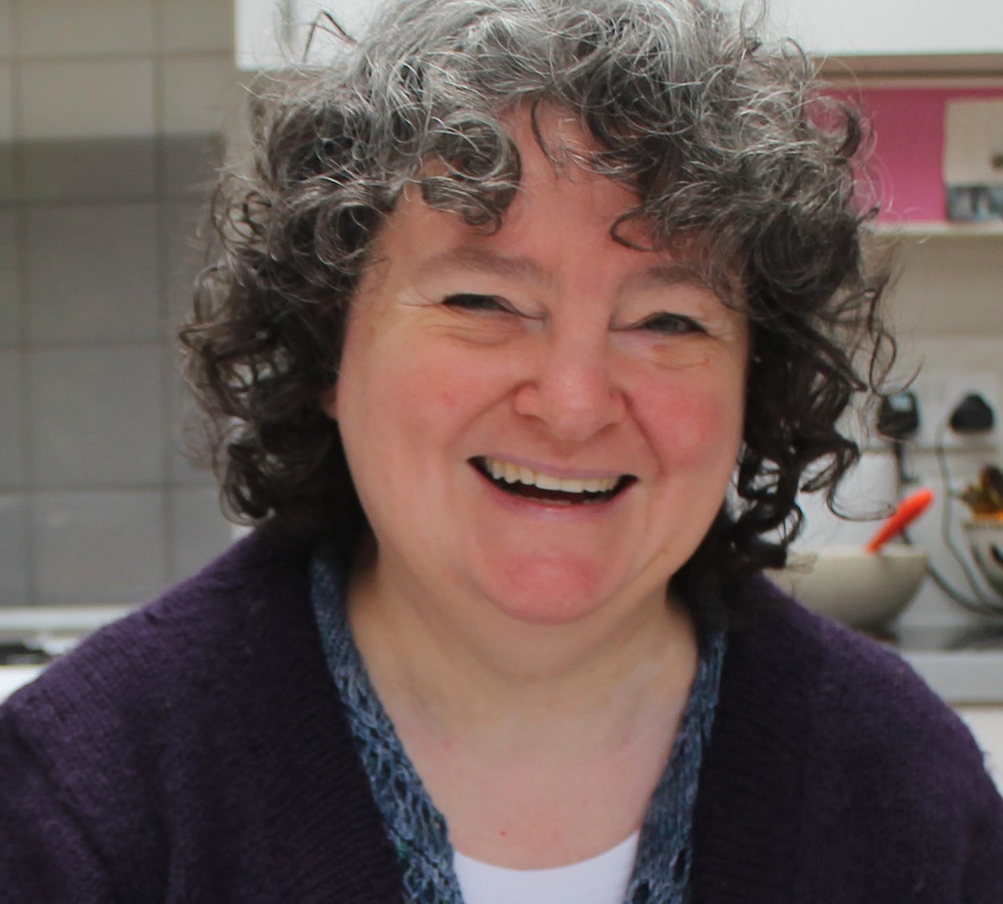 A photo of a laughing white women with dark curly hair. She is wearing in purple handknit cardigan and is sitting in a very nice kitchen. 