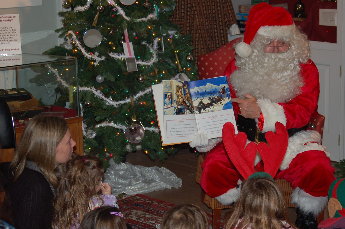 Color photograph of Santa Claus reading twas the night before christmas to a group of children in front of a christmas tree
