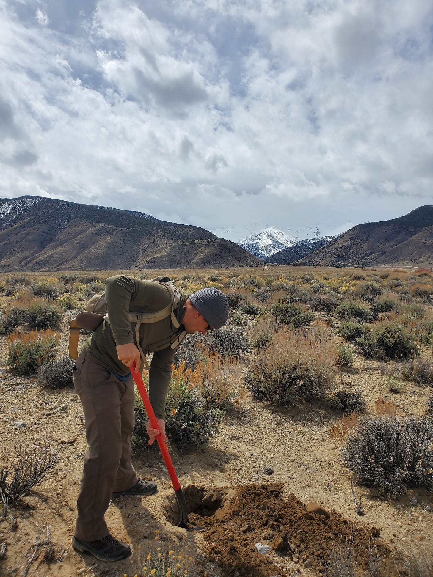 Eric digging in the ground with a shovel. Hills and White Mountains in distance behind him.