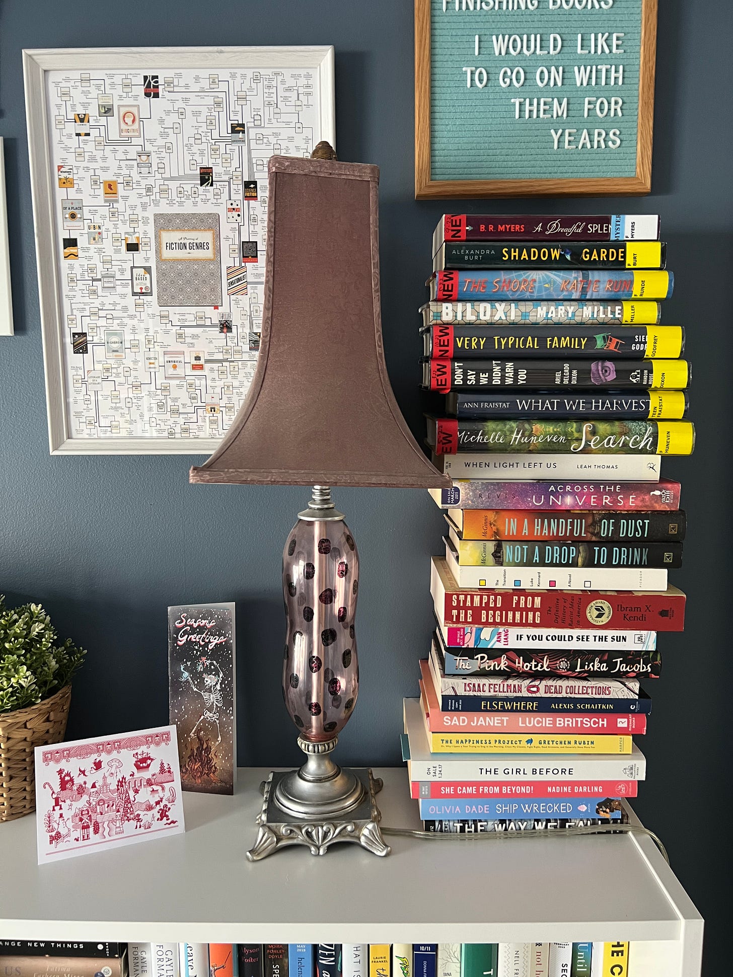 Gigantic book stack of library books and my own books on a shelf next to a lamp. 