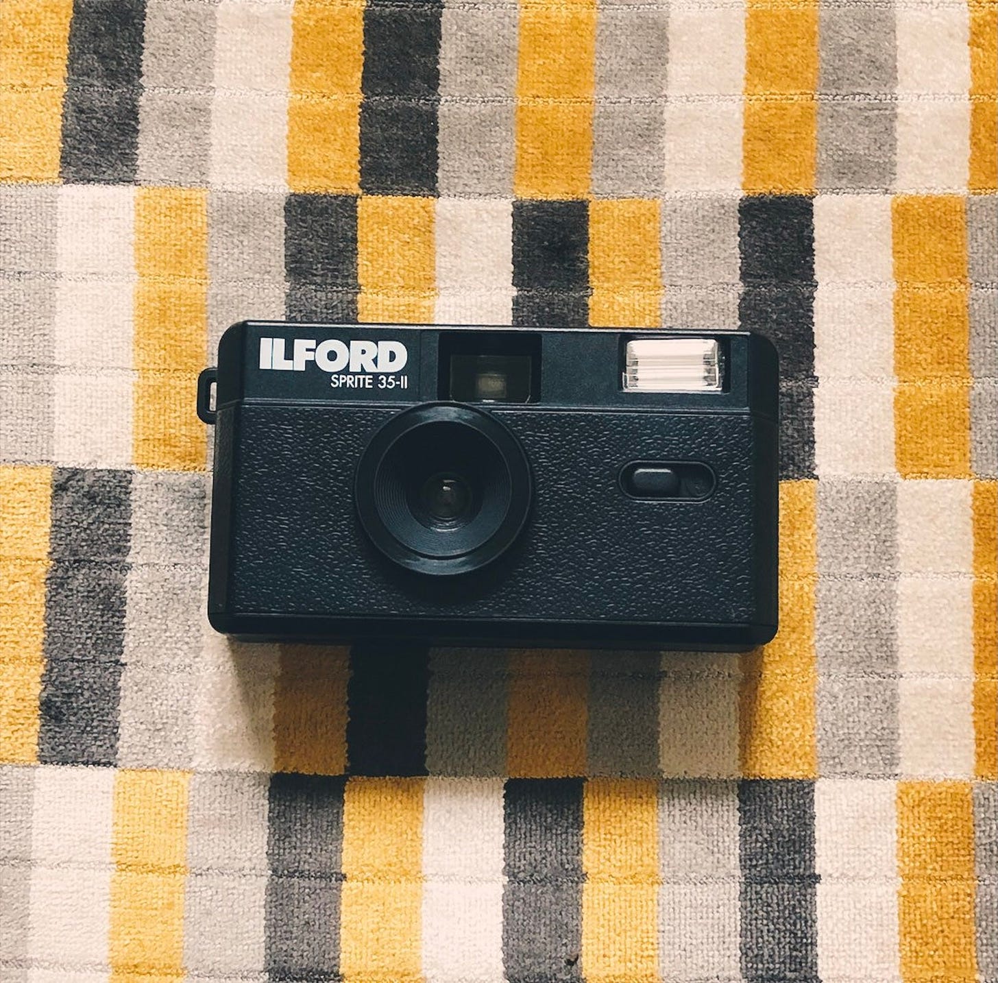 A black rectangular film camera with the word ILFORD in the top right corner in front of a striped carpet.