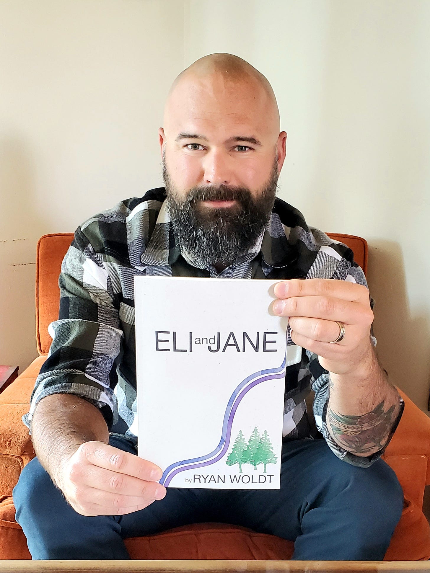 Eli & Jane book held in front of the camera by author Ryan Woldt wearing a black and white flannel shirt sitting on an orange chair. He has a bald head and a black and grey beard.