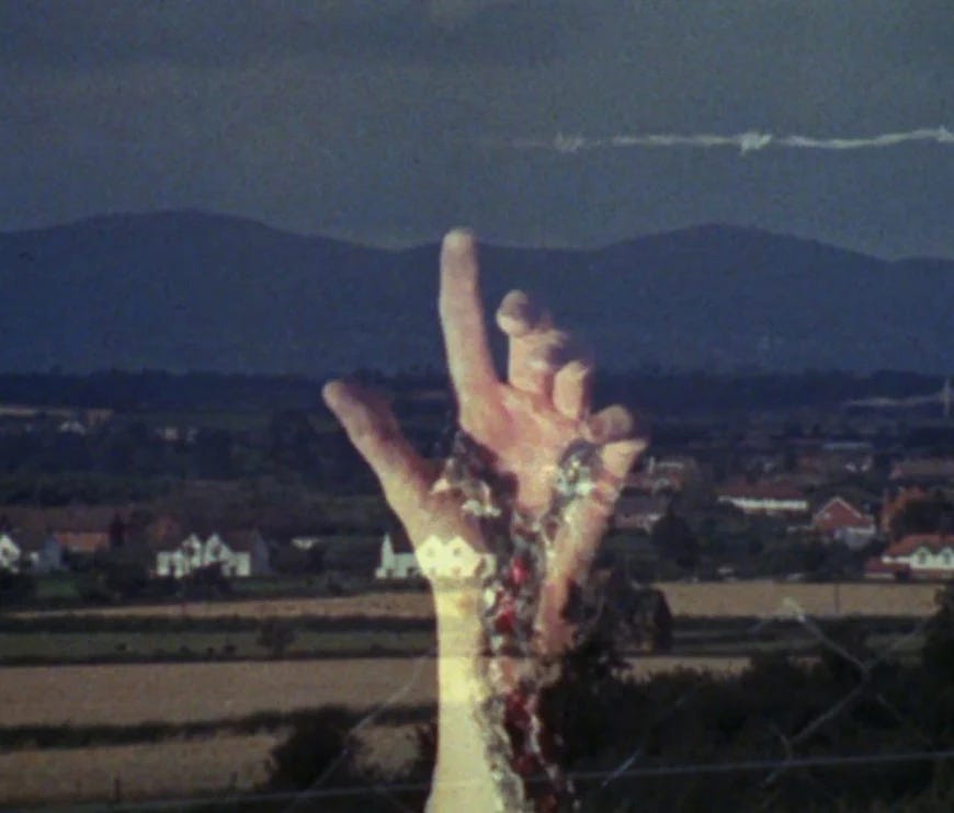 Still image from Penda's Fen, shot on a film camera, depicting a rural landscape with a hand in the foreground pointing upwards covered in scars.