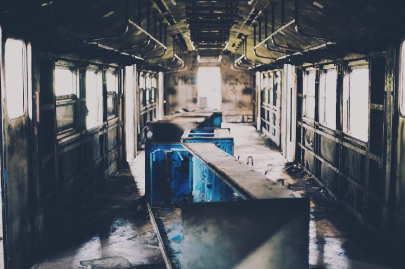 Uma foto de um vagão de um trem, abandonado e descascando. Simétrico, a perspectiva fica como um túnel. No primeiro plano, bem escuro; no fundo, bem iluminado. No meio do caminho, bancos tombados e azuis. Tudo muito sujo.
