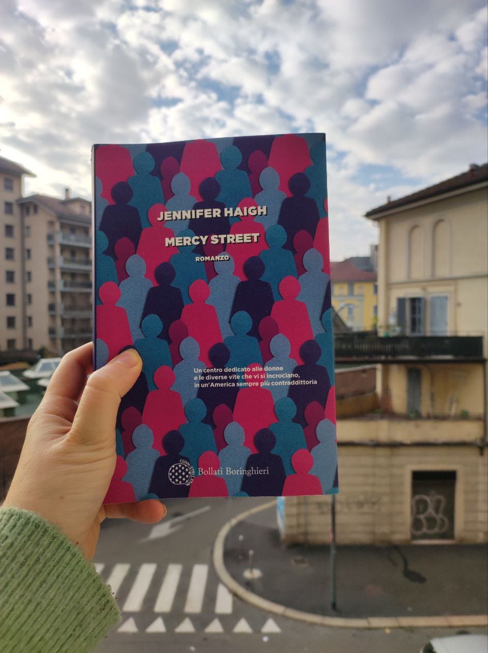 Una mano tiene un libro, dai colori blu viola e rosa. Il paesaggio di sfondo è la città di Milano, con condomini e la strada
