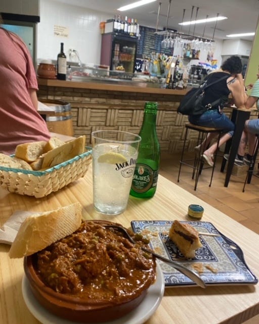 Photo of a brown earthenware bowl full of pork stew, with a piece of bread balanced on the side, on a table in a casual restaurant