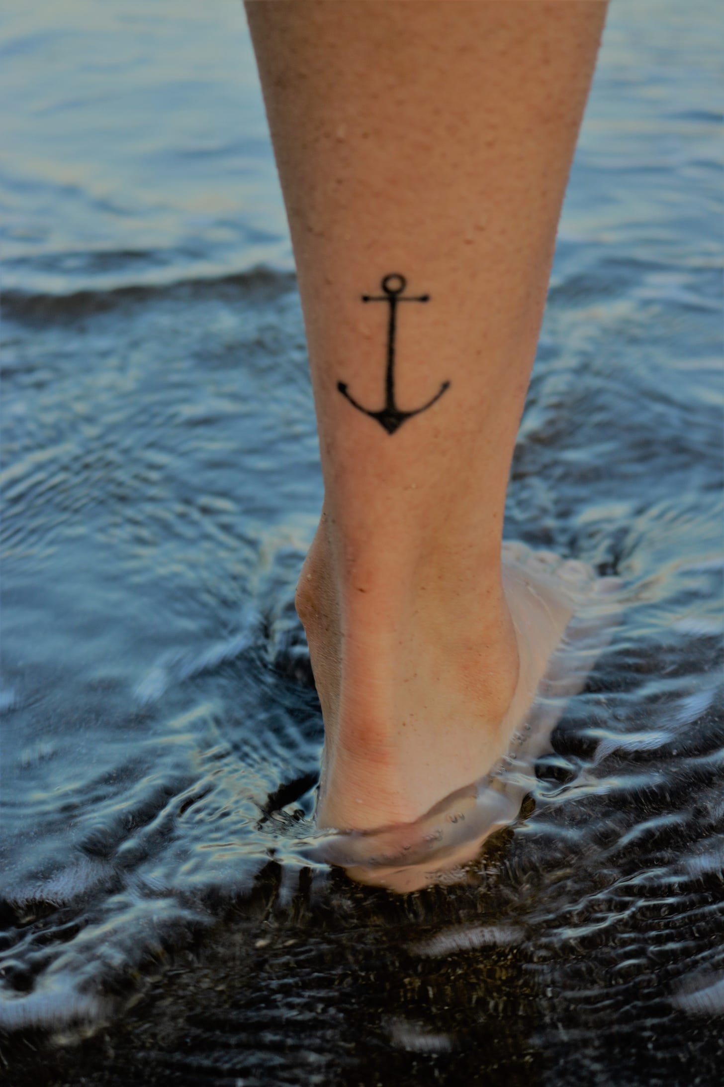 Back of a human calf above the Achilles tendon sporting a one dimensional anchor tattoo,standing in shallow water