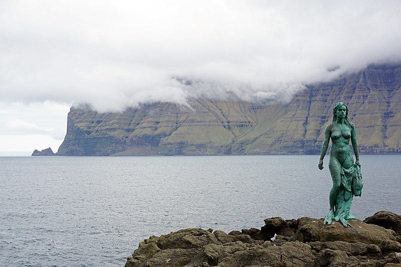 In the lower right corner, a bronze (bluey green) statue of a naked woman with long hair is holding a seal skin in her left hand. She is facing the camera and standing on a rock. In the background, thick white clouds hang over a green rocky land mass. The sea is between both of them.