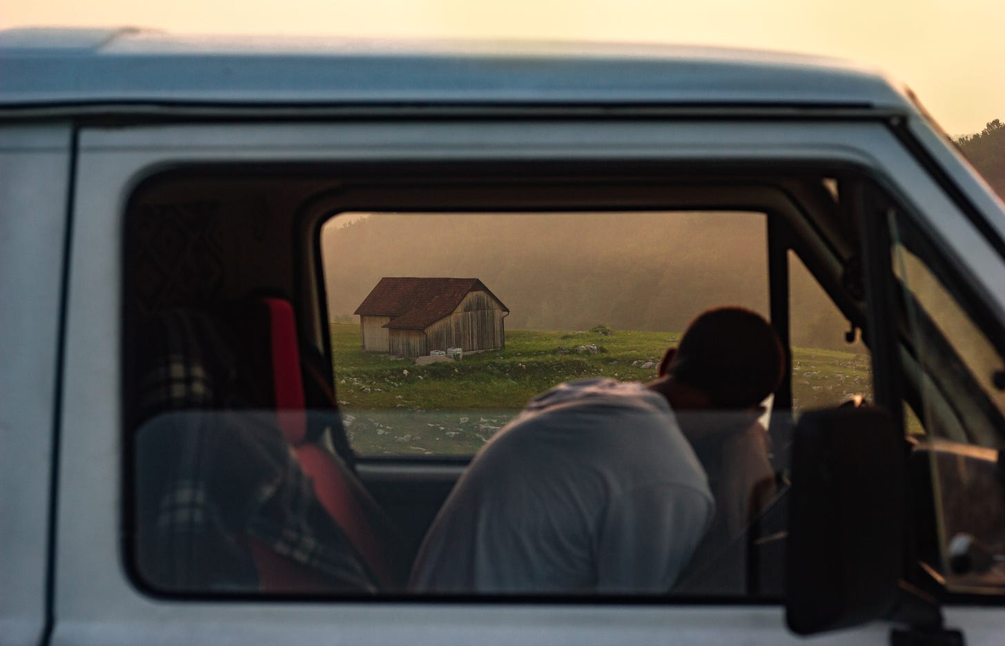 A close up of the window of a van, a man lying his head on the wheel. In the background is a lonely small wooden cabin surrounded by stunning scenery.