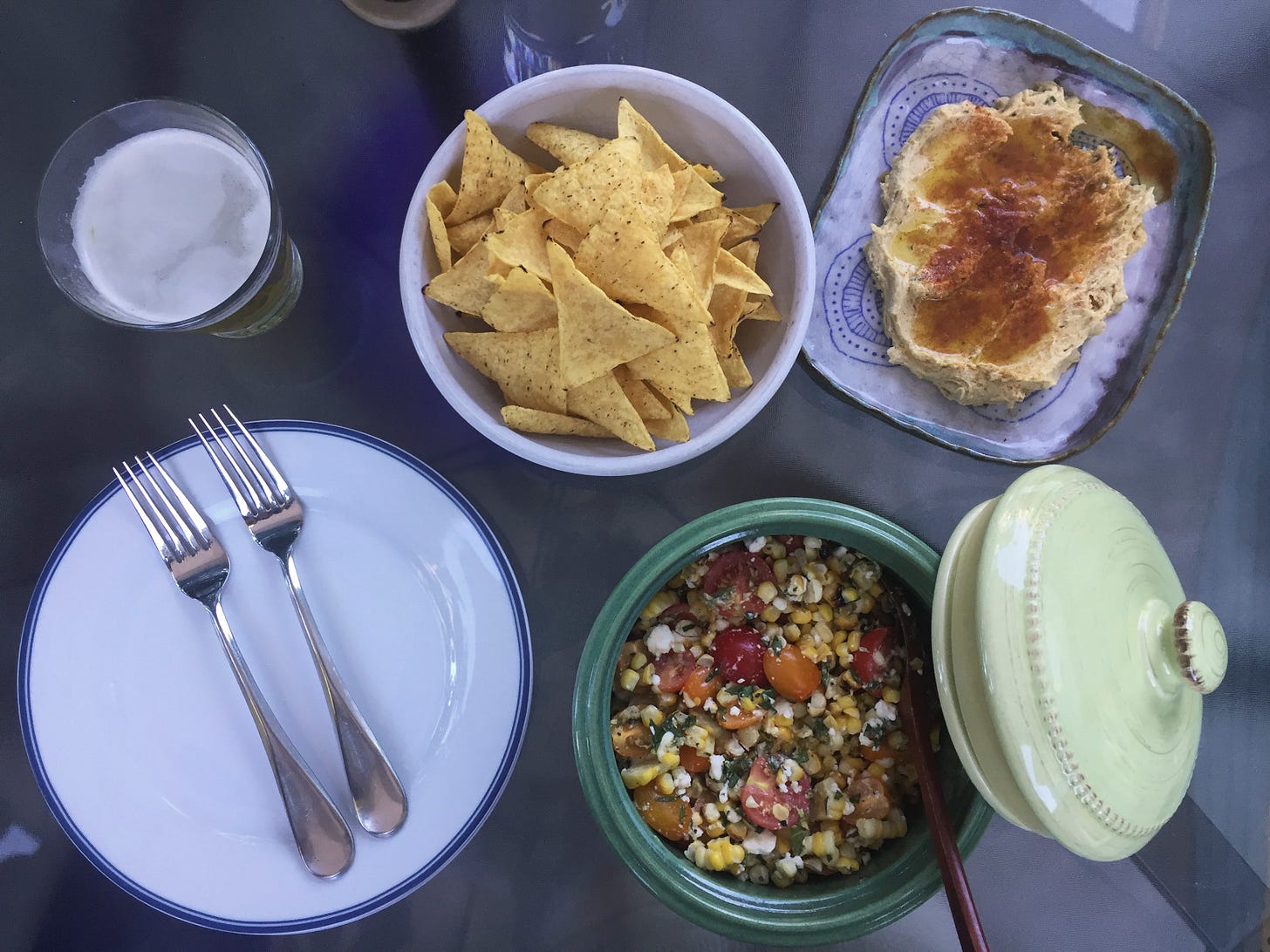 An arrangement of serving dishes with tortilla chips, the corn salad, and hummus with oil and spices on top. A plate and a glass of beer are in the left side of the frame.