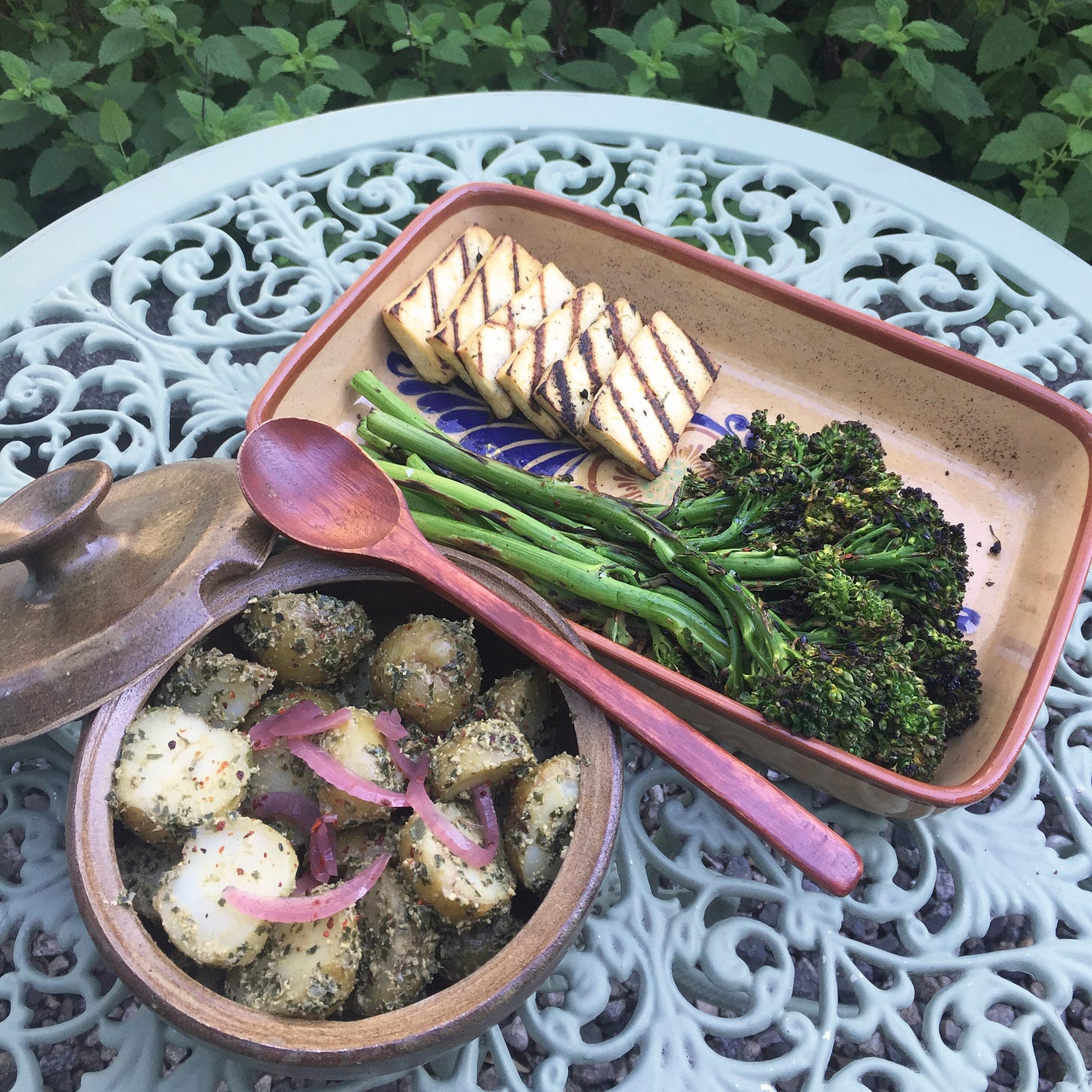 On a wrought-iron outdoor table, two stone dishes: a round tureen with pesto potato salad with pickled onion on top, and a rectangular shallow dish with slices of grilled tofu and long pieces of charred broccolini. A spoon rests at the edge of the potatoes.