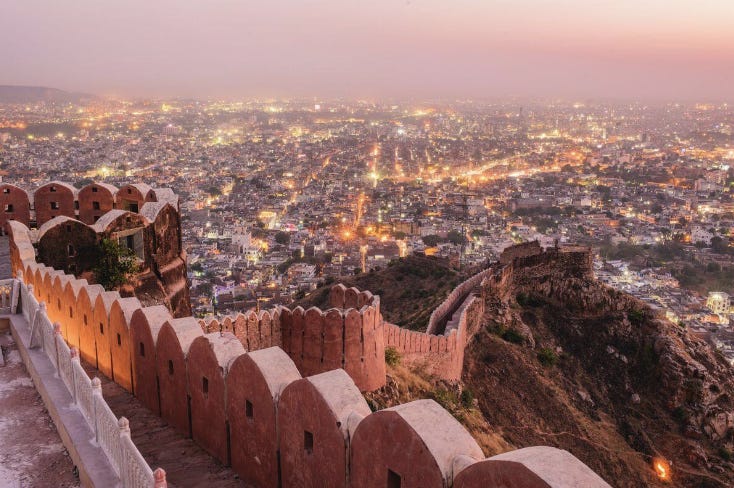 A city in the dusk light filled with pink buildings. Some of the buildings are light up with golden lights. The sky is pink in the dusk light. In the foreground of the picture is a pink stone wall of a fort running along the mountain cliff.