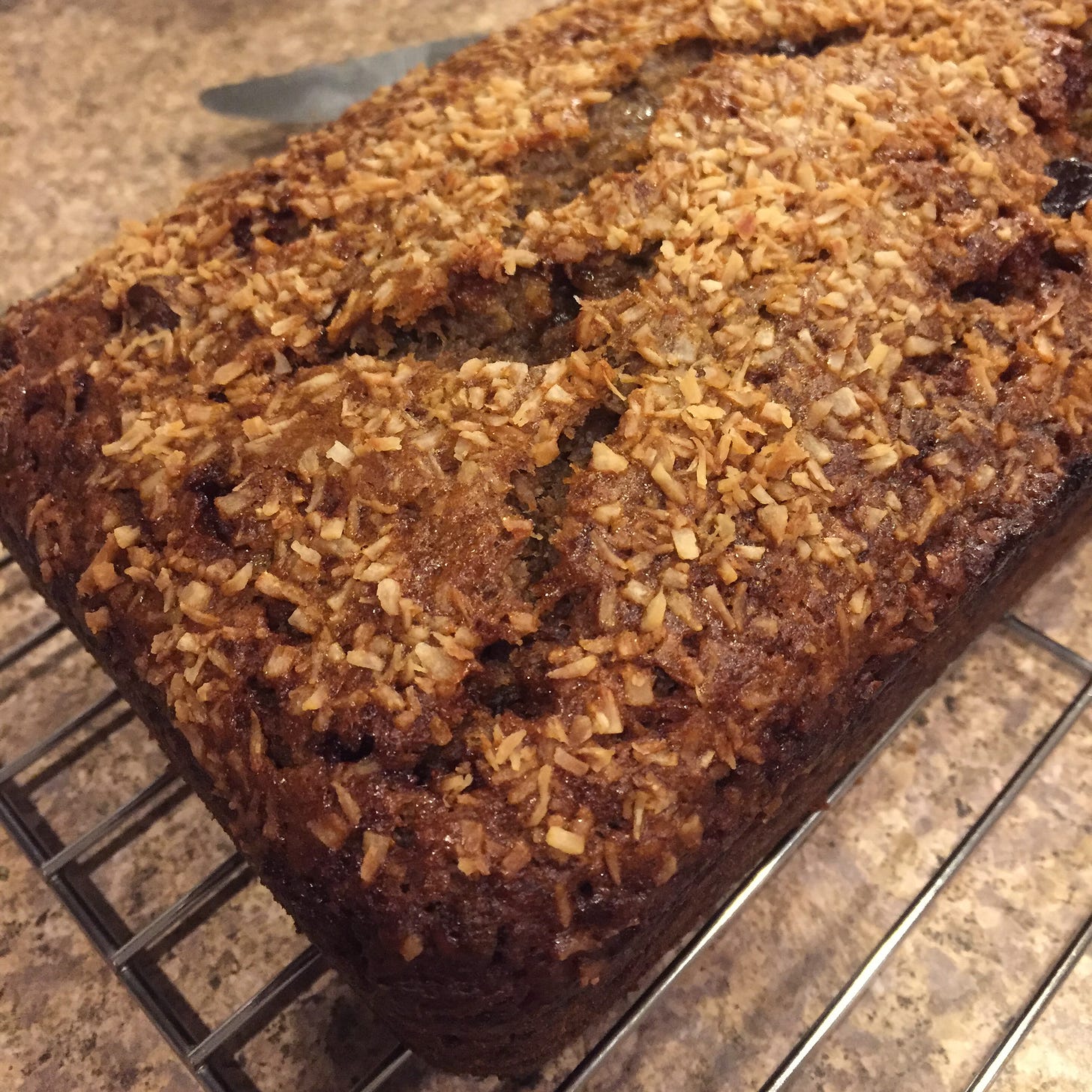 On a cooling rack, a brown loaf of banana bread with toasted coconut and a top that has cracked in places.