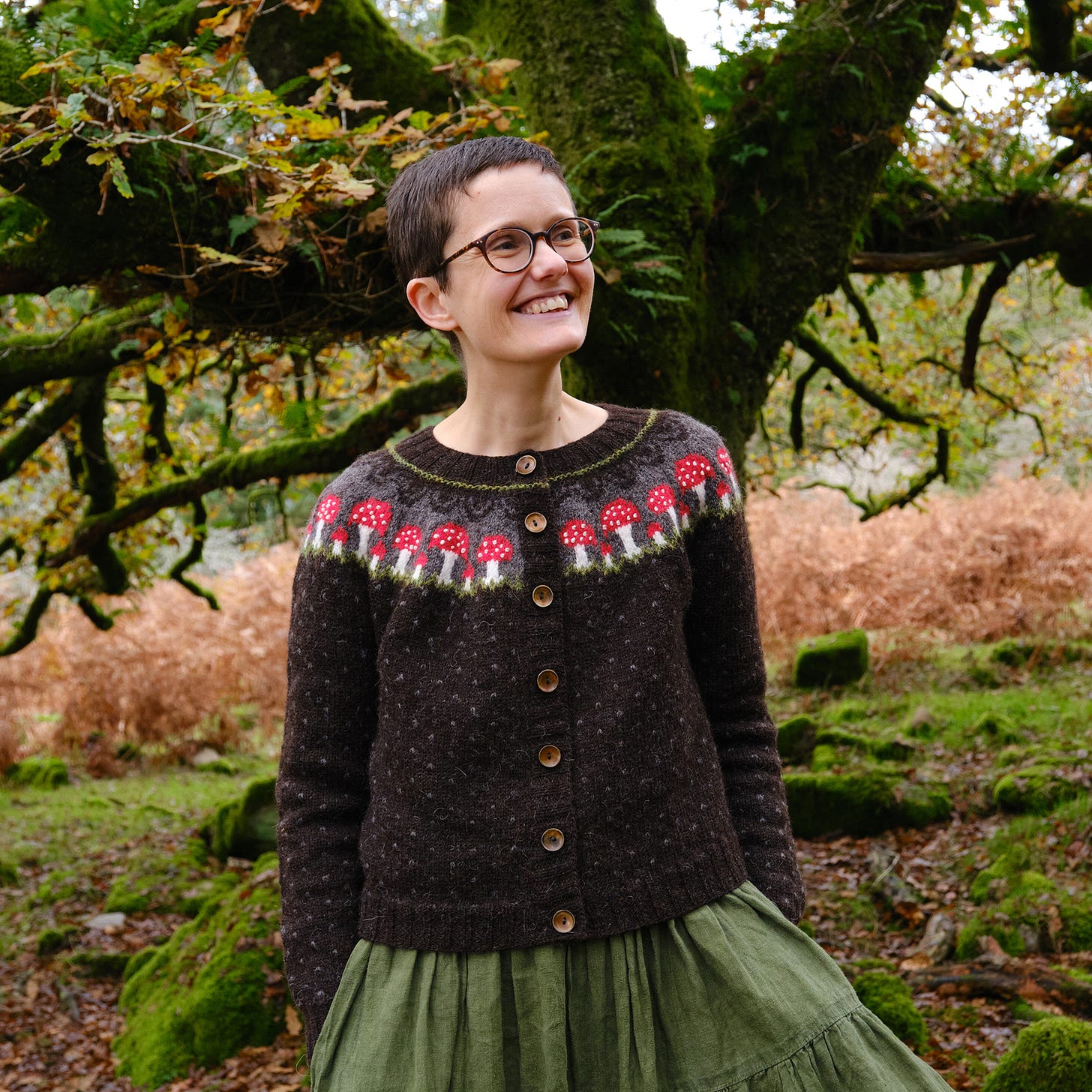 Image description: Katie, a white human with short hair and glasses, with hands in pockets and looking off to right of frame, grinning. Katie is wearing a dark brown spotted cardigan with a colourwork yoke of red spotty fly agaric mushrooms. Behind Katie is a big mossy oak tree. 