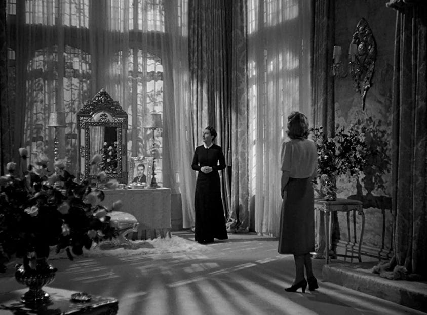 Judith Anderson as Mrs. Danvers in Rebecca (1940) stands in front of a draped window. Joan Fontaine as the second Mrs. de Winters looks at her. 