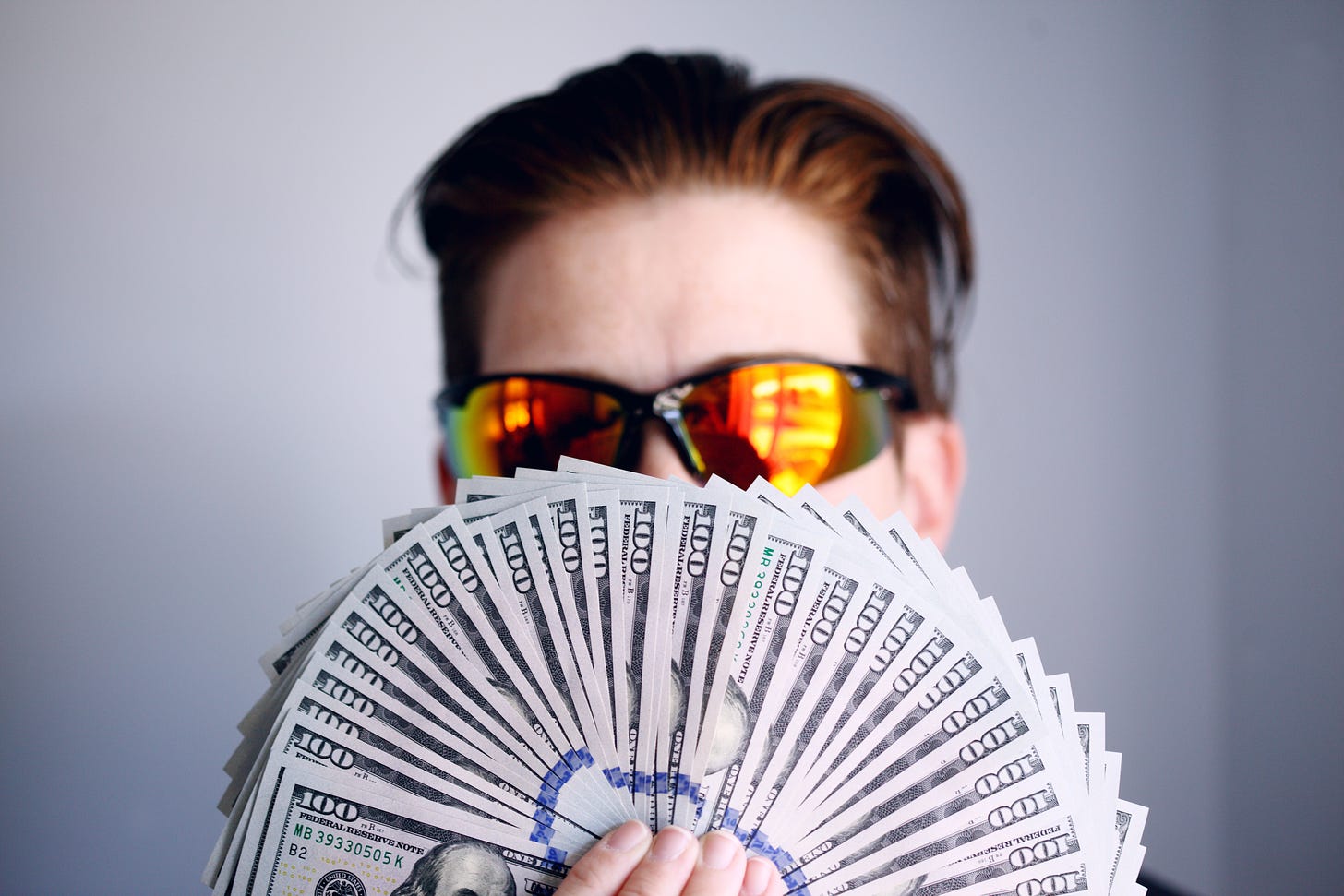 person wearing sunglasses, hiding behind a fan of one hundred dollar bills