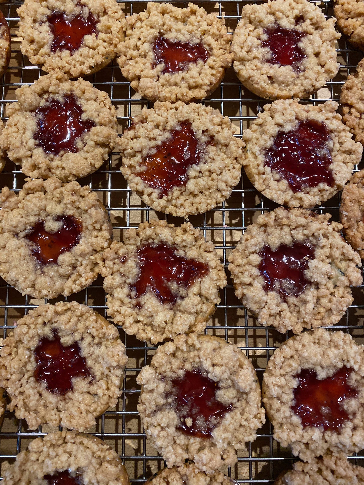 Three rows of round cookies on a metal cooking rack. The cookies have a crumbly streusel topping, and the centers are filled with raspberry jam, which is glistening and bubbly from being in the oven.