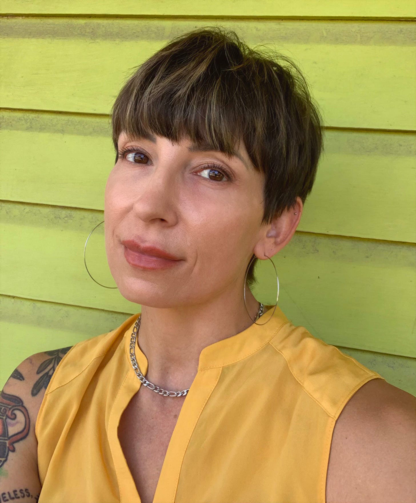 A nonbinary person in a yellow tank top leans against green house shingles. They wear hoop earrings and a short haircut about their ears