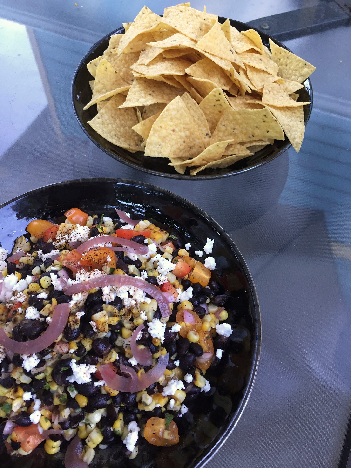 Two shallow black bowls on an outdoor table; one in the background has yellow corn tortilla chips. The one in the foreground has a black bean and corn salad with pieces of tomato and red pepper, and overtop are crumbles of feta and slices of pickled red onion.
