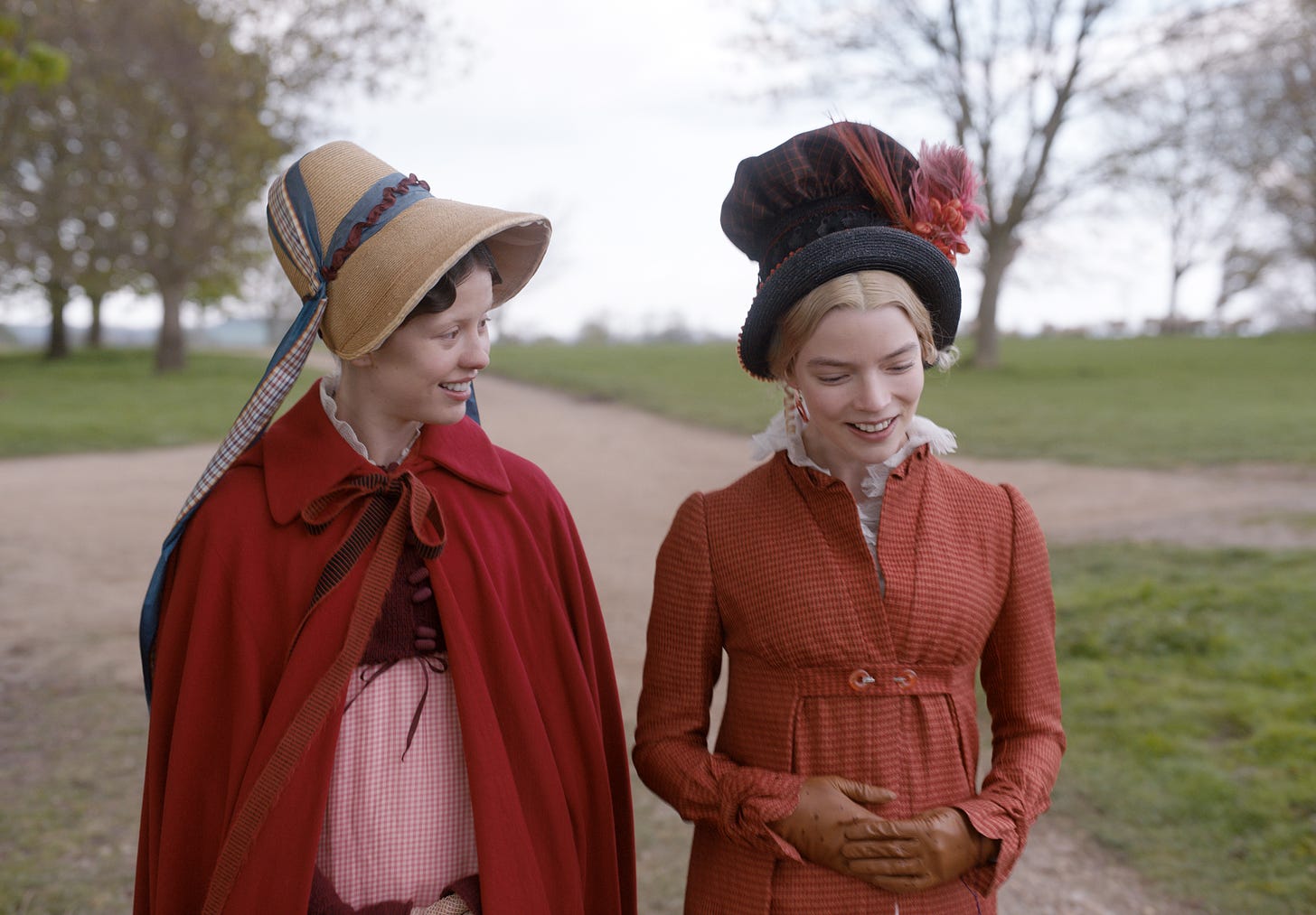 Actors Mia Goth and Anya Taylor-Joy are in rust-red Regency costumes, wallking in the countryside and talking, as they depict the characters of Harriet Smith and Emma Woodhouse in Autumn de Willde's 2020 film adaptation of 'EMMA.' 