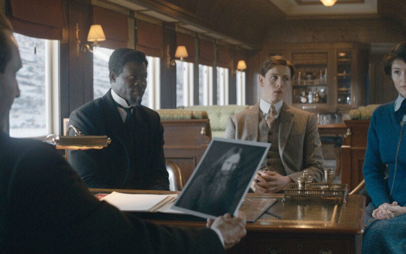 The Duke, Shola, Conrad, and Polly seated around a desk on a train. The Duke looks over a picture of Rasputin.