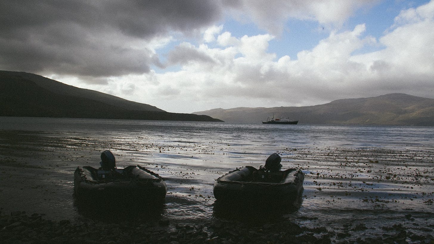 A photograph of a harbour, with a beach in the foreground with two zodiac boats ashore, glassy dark water goes out to a ship far away in the backrgound, and behind that hills and dark clouds.