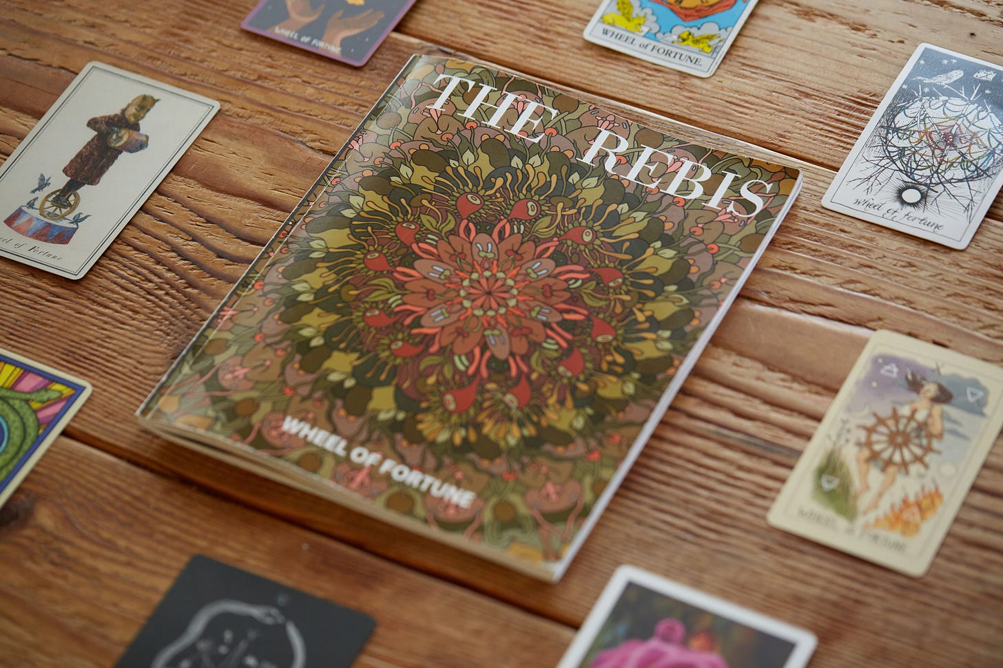 The Rebis: Wheel of Fortune, a magazine featuring an earth-toned mandala with greens, reds, and brown elements, sits at the center of a circle of tarot cards from various decks. Every tarot card is a different interpretation of the Wheel of Fortune.