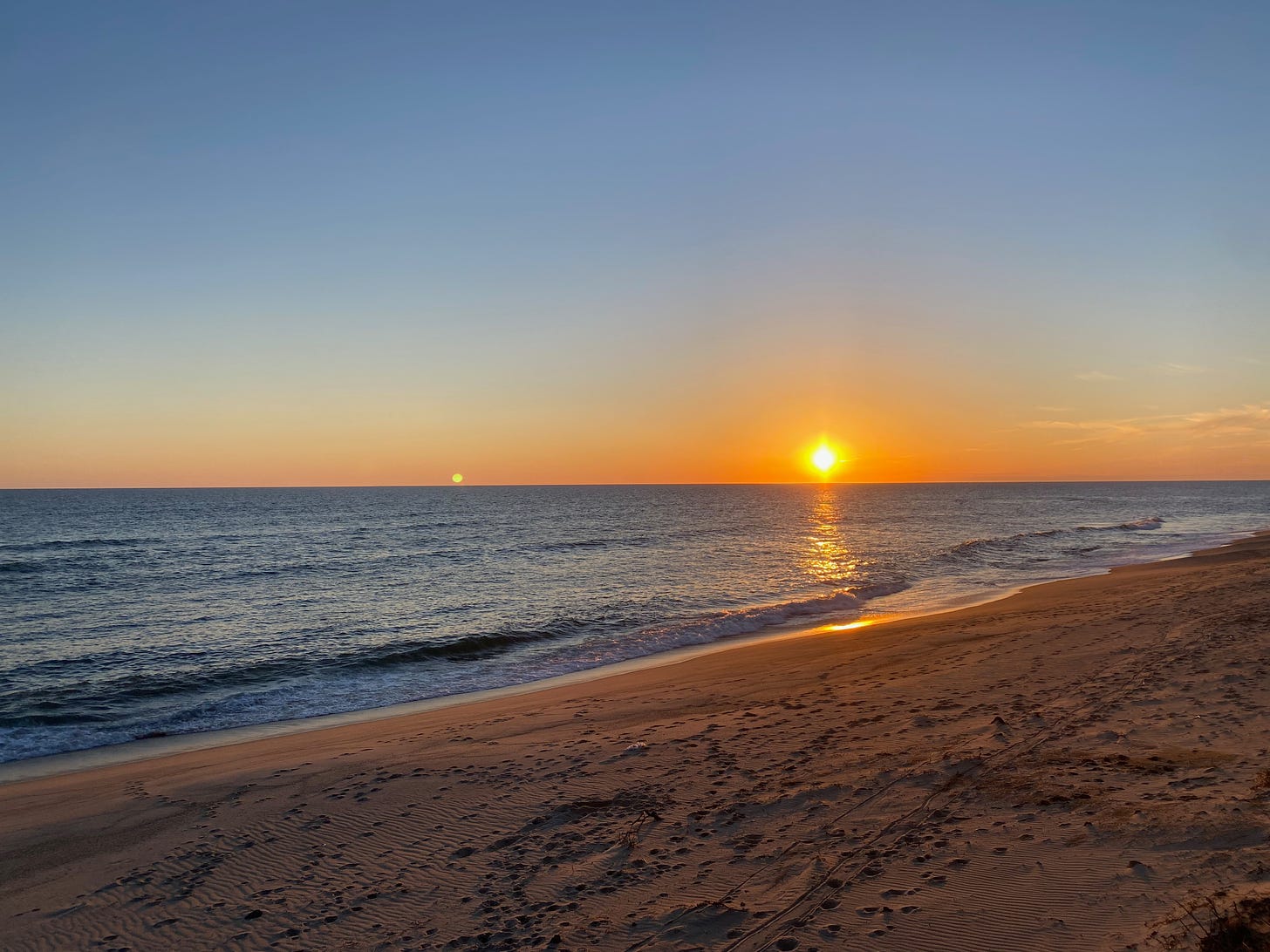 A beach at sunset. The ocean is still, the sky a clear, deep blue, and the golden sun is low on the horizon, reflecting on the ocean and in the breaking waves.