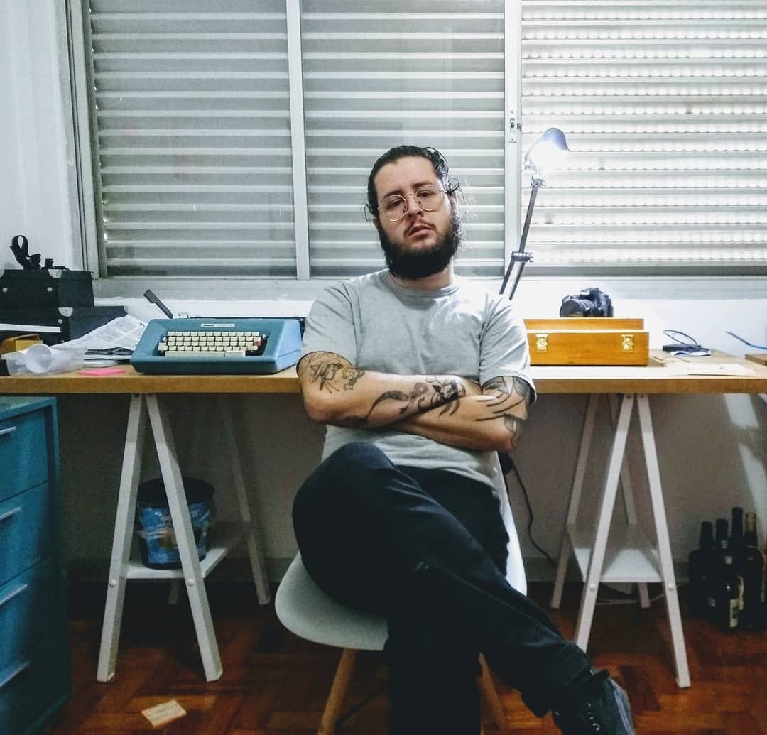 Angelo sitting in a chair in front of a table with a typewriter and other things, like a camera, a wooden box, some paper and boxes