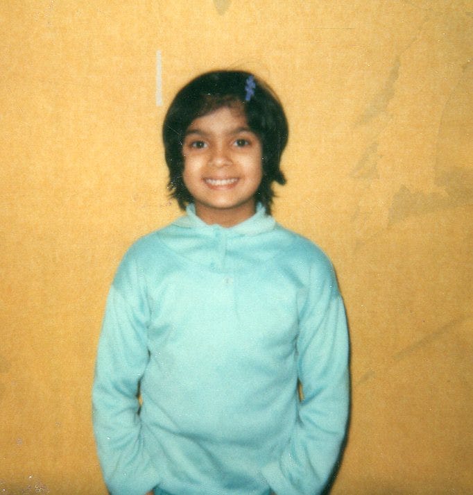 A picture of Nisha, age 6 or 7. She is standing against a yellow backgrop. She has a light blue pullover. Her hair is black and chin-length and she has a matching barette in her hair.